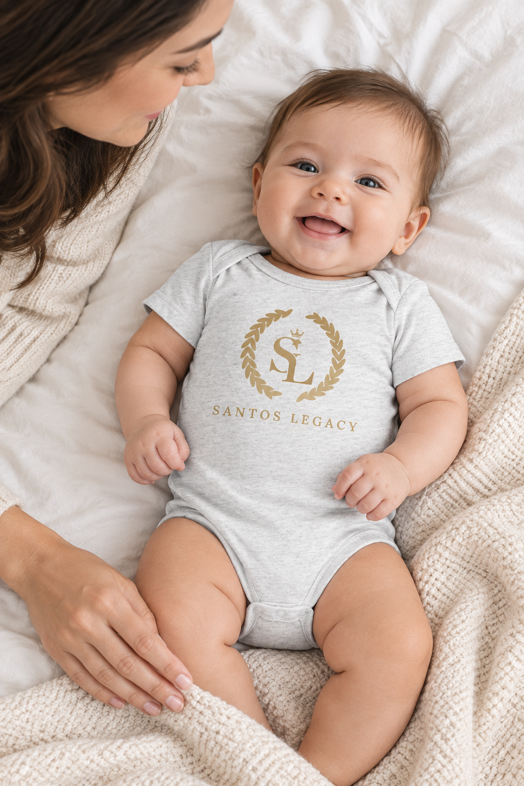 Mother smoothing a blanket beside a baby wearing the Santos Legacy Little Legacy Baby jersey bodysuit on a bed