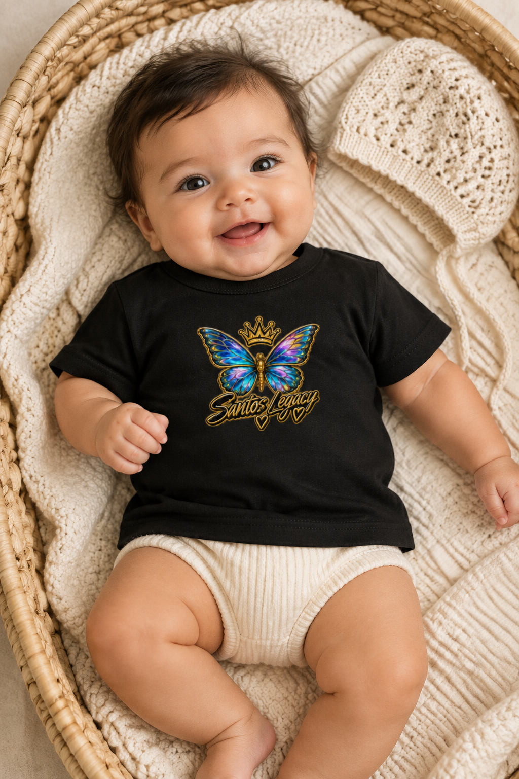 Baby wearing the Santos Legacy Baby Jersey Short Sleeve Tee lying on a woven basket bassinet with a knitted bonnet