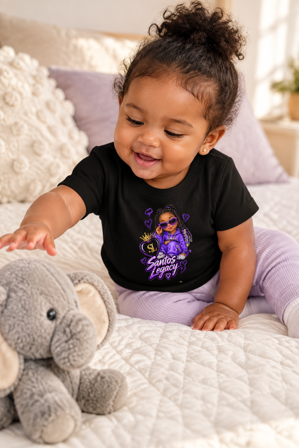 Baby wearing the Santos Legacy Baby Jersey Short Sleeve Tee on a quilted bed reaching for a plush elephant toy