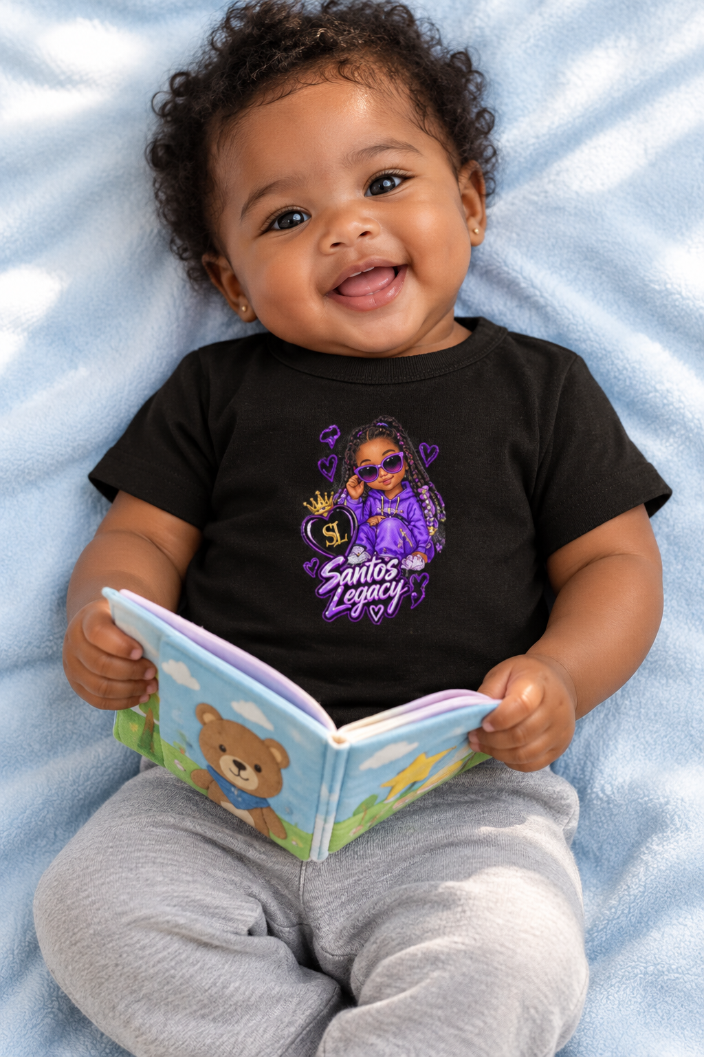 Baby wearing the Santos Legacy Baby Jersey Short Sleeve Tee holding a soft story book on a pale blue blanket