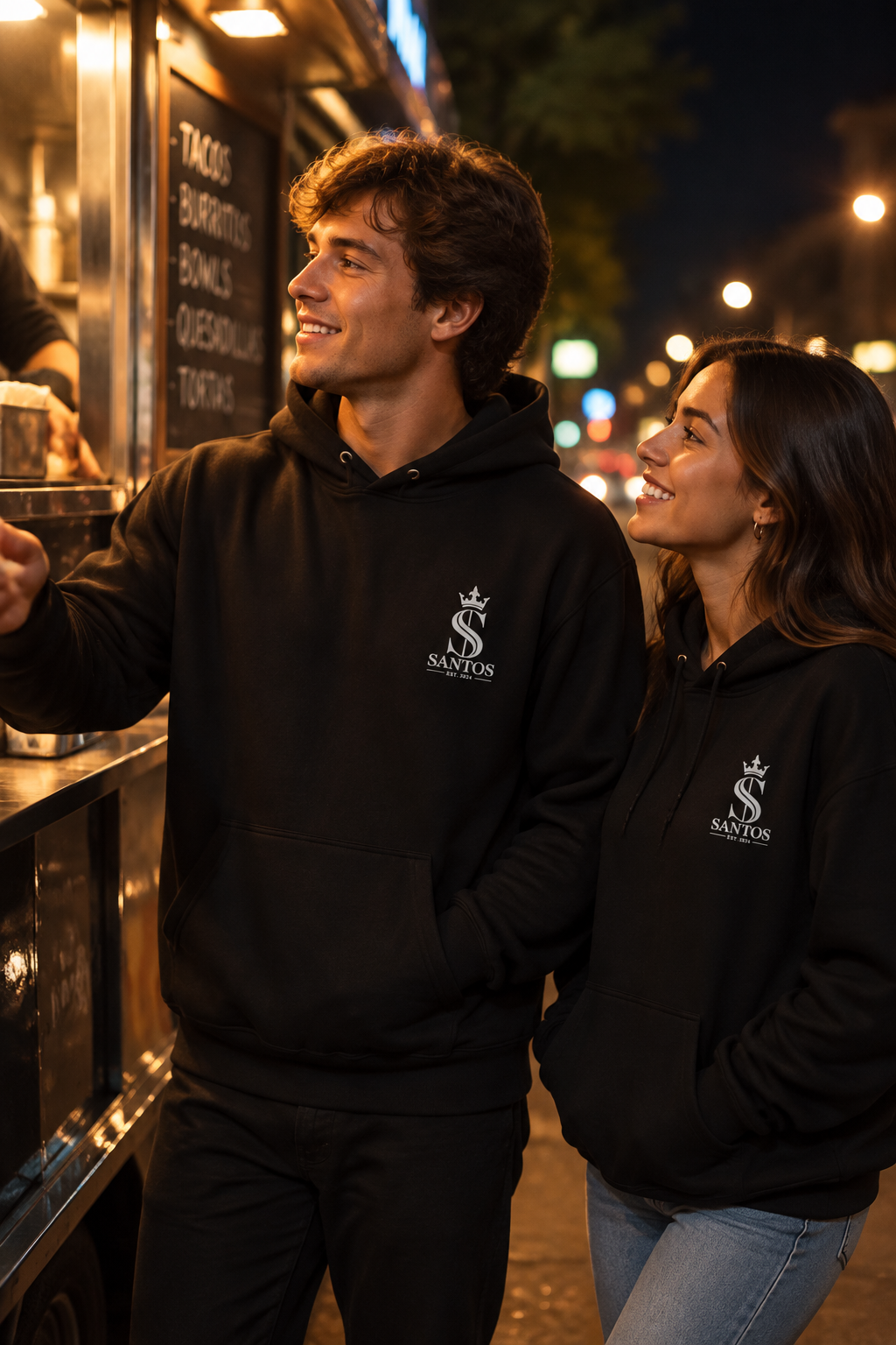 Couple wearing the Santos Legacy Signature Premium Unisex Hoodie ordering at a food truck at night